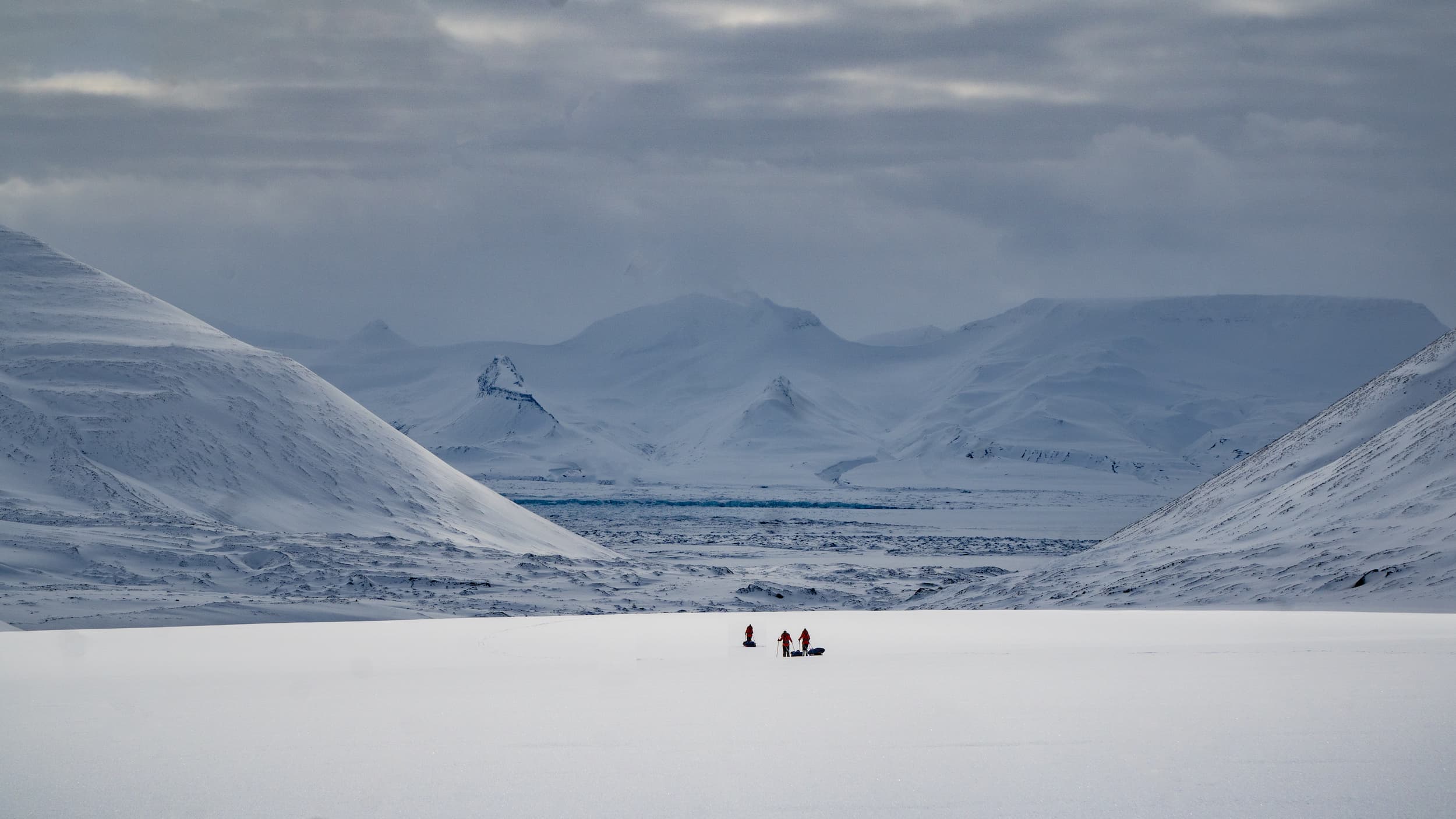 Snow-covered mountains surround a vast white plain where three small figures traverse the icy landscape on sleds or skis.