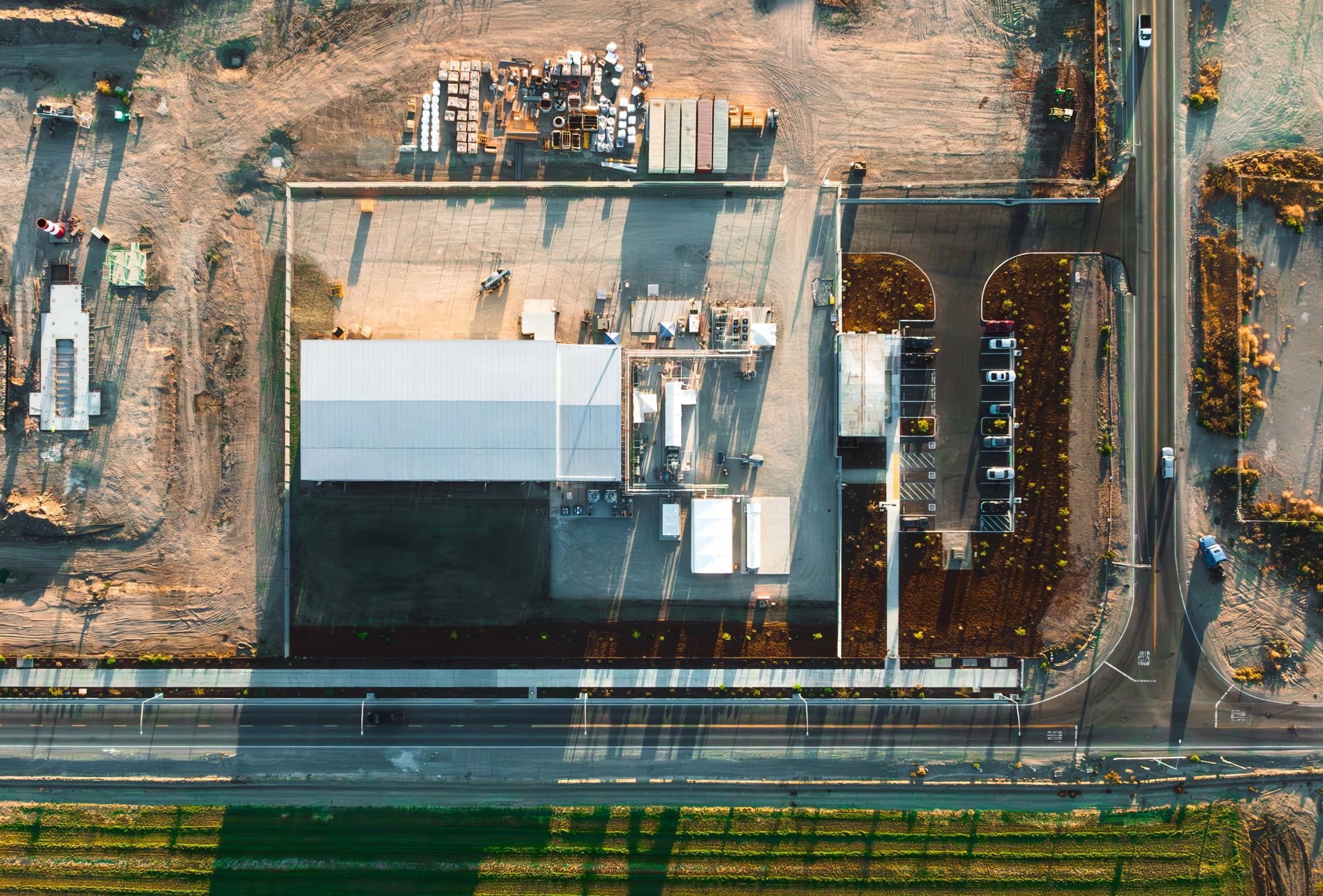 Aerial view of an industrial site with buildings, equipment, and storage areas next to a road and green field.