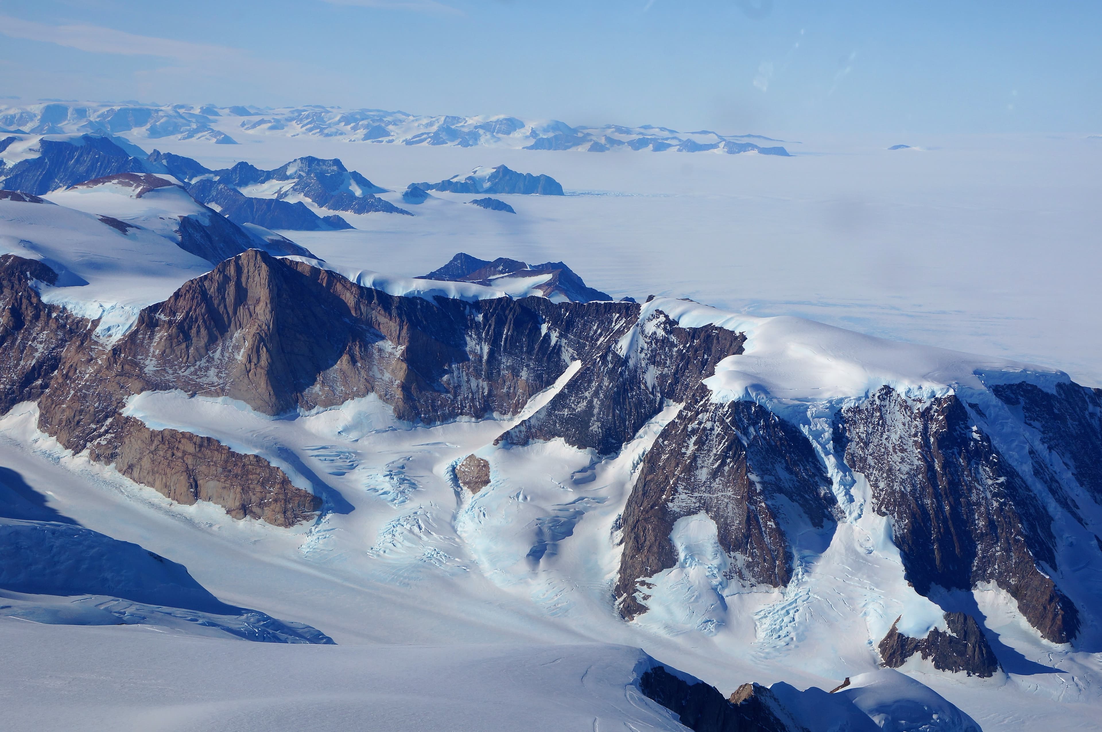 Aerial view of snow-covered mountain peaks and glaciers in a rugged, icy landscape surrounded by clouds and distant mountains.