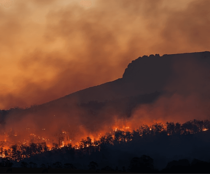 Raging wildfire engulfs forest at mountain base, orange smoke billows against dark silhouette of cliff under fiery sky.